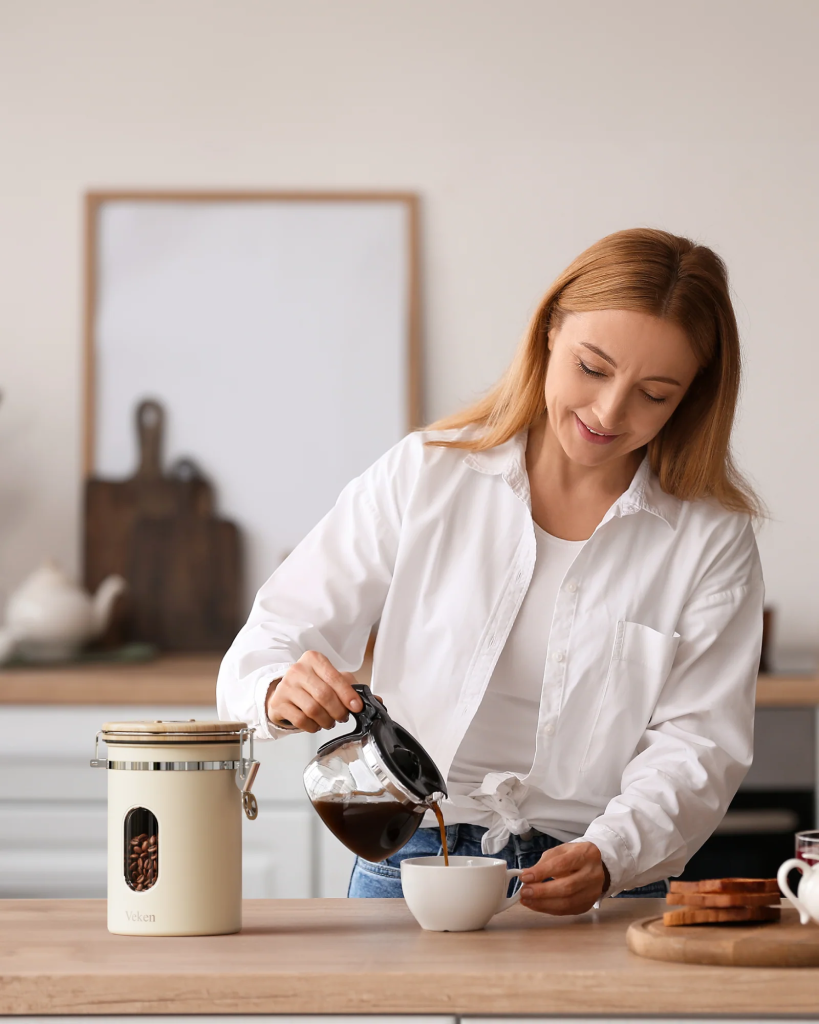 Female with auburn hair wearing a cotton white long sleeve button up shirt pouring a cup of coffee with cream color Veken coffee canister next to her